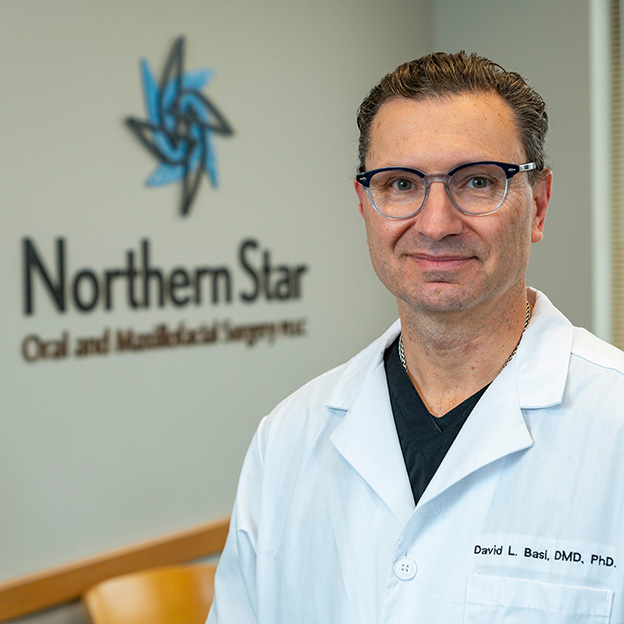 The image shows a man wearing glasses, standing in front of a sign with the text  Northern Star,  smiling at the camera. He appears to be a professional, possibly a dentist or medical practitioner, given his attire and the context provided by the sign behind him.