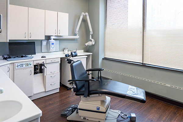 The image shows an interior view of a dental office with various pieces of equipment and furniture, including a dental chair, examination table, sink, cabinet, and other medical supplies.