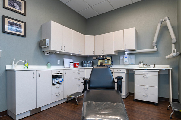 The image shows a well-equipped dental office with various pieces of equipment, including a chair, sinks, and cabinets, set against a dark wall.