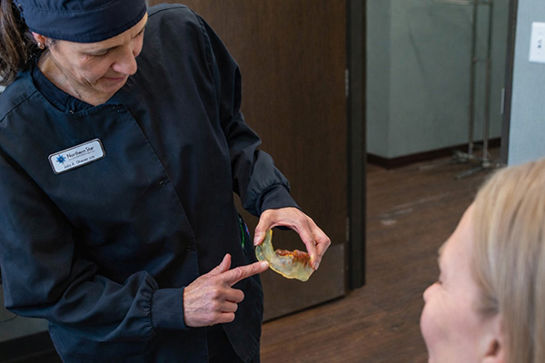 The image shows a woman standing behind a counter with a box of tissues, holding a tissue in her hand, wearing a white lab coat and a stethoscope around her neck, indicating that she is a medical professional, likely a nurse.